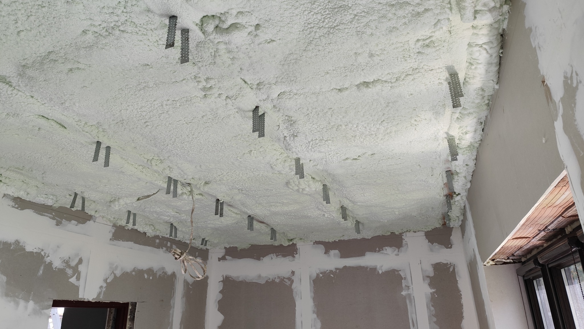 Ocieplenie pianką PUR sufitu z widocznymi metalowymi elementami mocującymi. Ściany w trakcie przygotowania do wykończenia, widoczne okno z roletą.