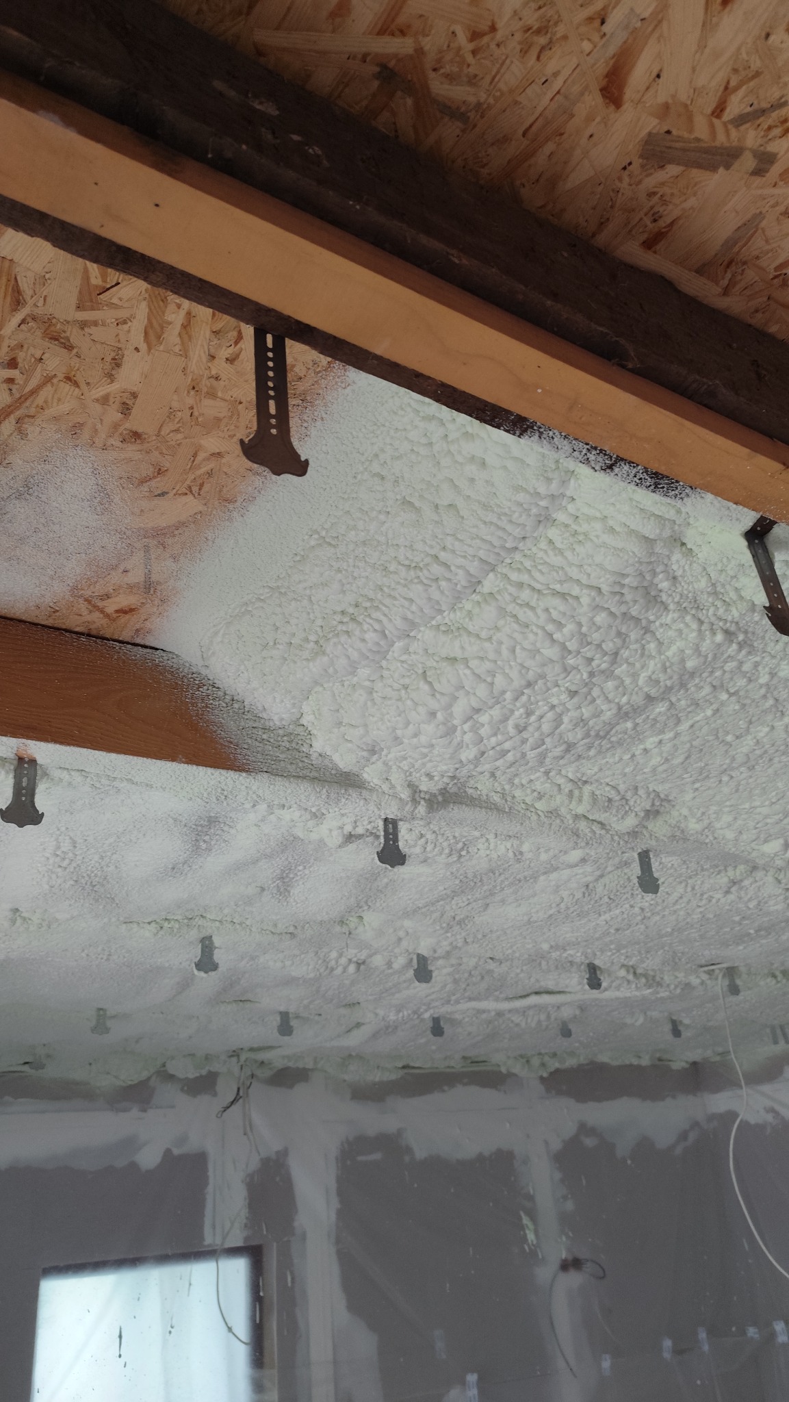 Ocieplenie poddasza pianką PUR, widoczne krokwie i metalowe uchwyty mocujące. Piana w kolorze białym, o nieregularnej strukturze, pokrywa całą powierzchnię.