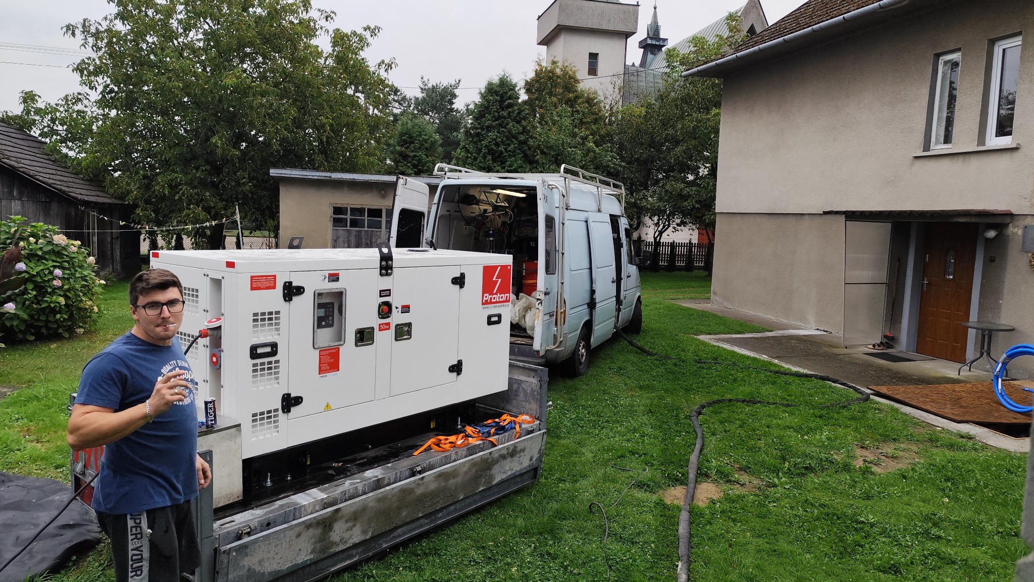 Agregat Proton w busie na trawie, widoczny mężczyzna z napojem, budynki w tle. Sprzęt gotowy do pracy na zewnątrz, zasilanie w trakcie realizacji.