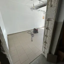 Świeżo ułożone, beżowe płytki podłogowe w garażu, widok zza surowej, betonowej framugi drzwiowej, z odkurzaczem przemysłowym w tle i automatyką bramy garażowej na suficie.