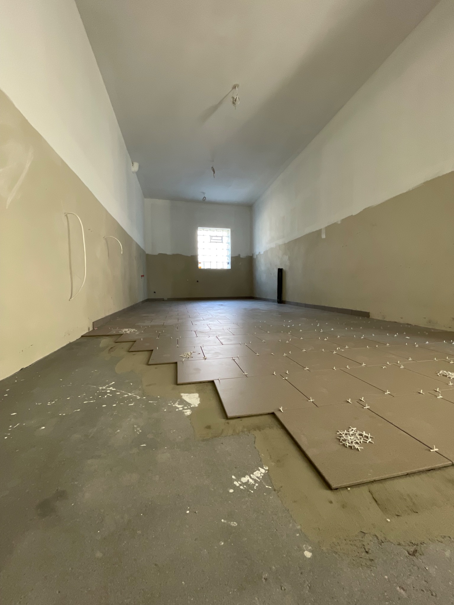 Układanie płytek podłogowych w długim, pustym pomieszczeniu z oknem w tle, widoczne krzyżyki dystansowe i ślady zaprawy.