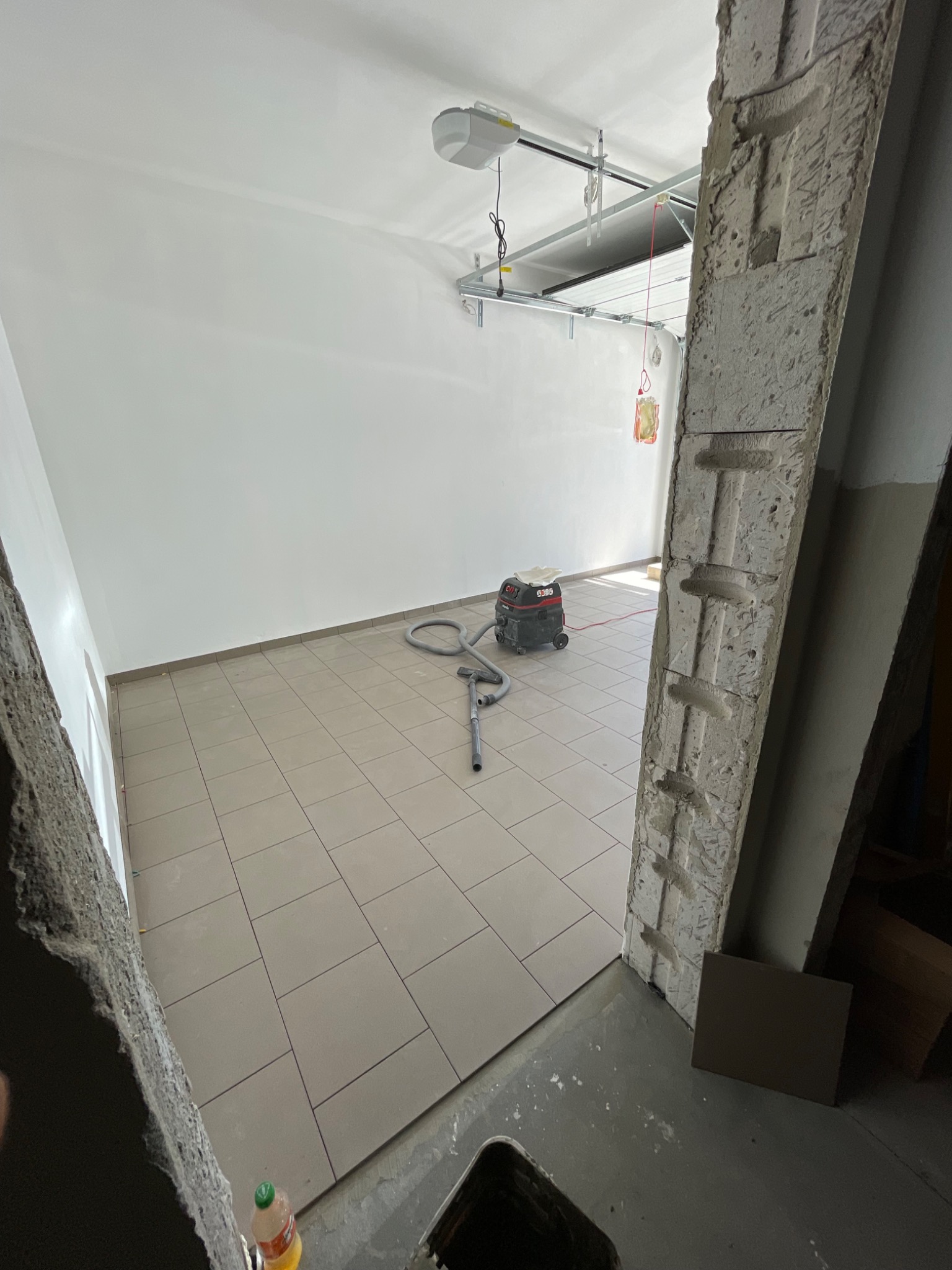 Świeżo ułożone, beżowe płytki podłogowe w garażu, widok zza surowej, betonowej framugi drzwiowej, z odkurzaczem przemysłowym w tle i automatyką bramy garażowej na suficie.