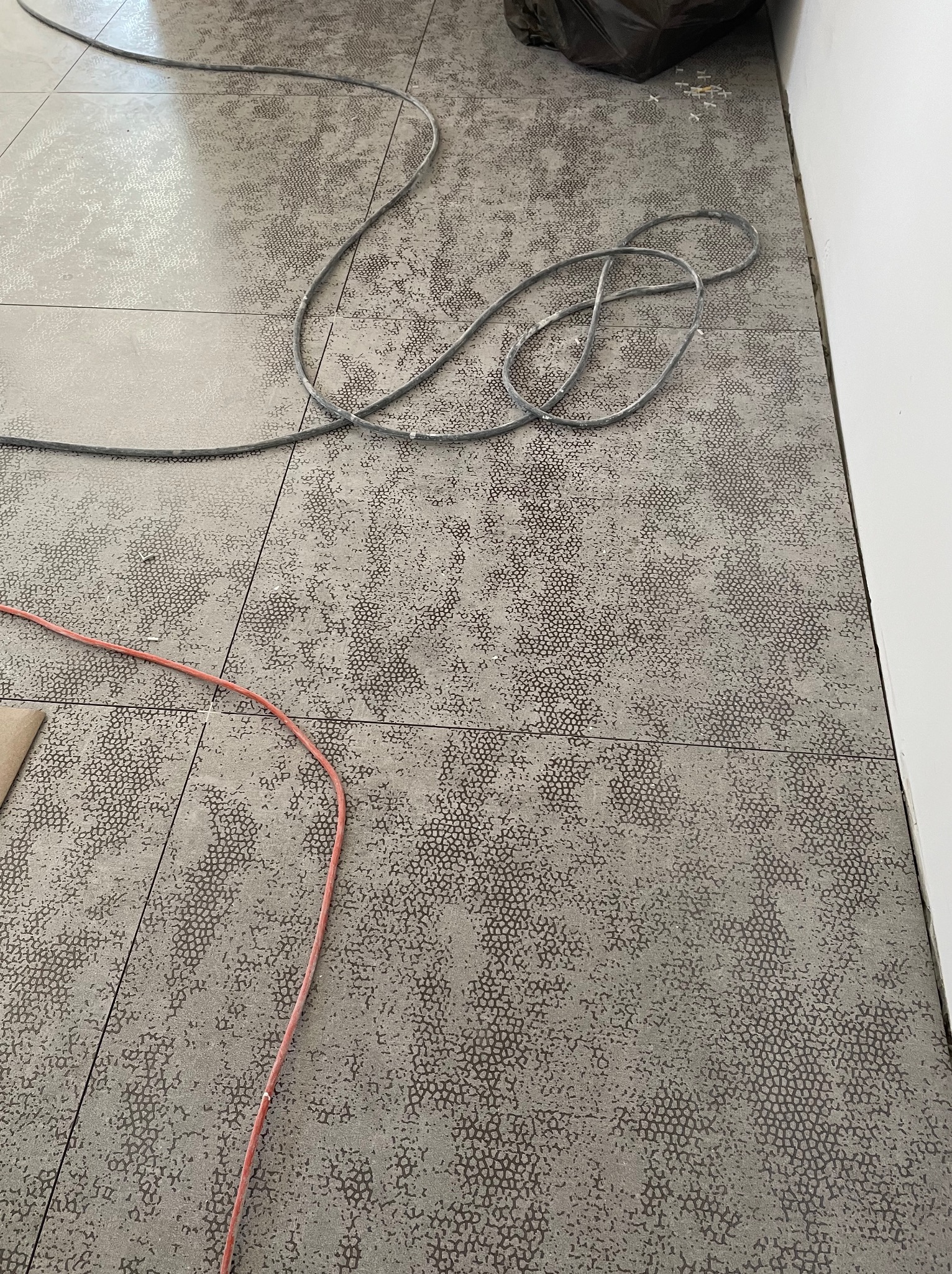 Szare płytki podłogowe z wzorem w trakcie układania, widoczne kable i worek na śmieci w rogu.