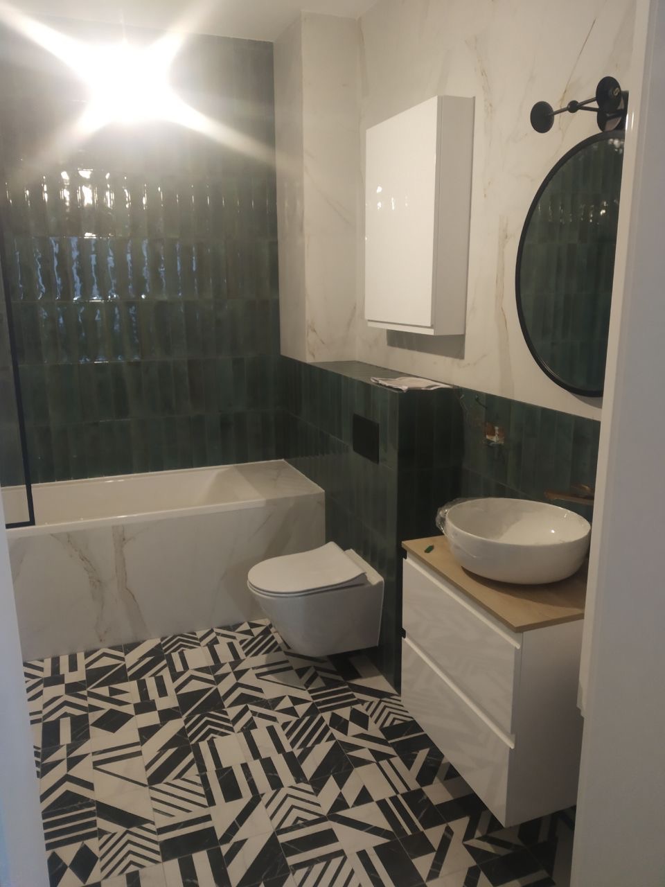 Wykończona łazienka z zielonymi, pionowo ułożonymi płytkami przypominającymi cegiełki, geometryczną podłogą w czarno-białe wzory, białą umywalką nablatową i szafką z jasnym drewnianym blatem.