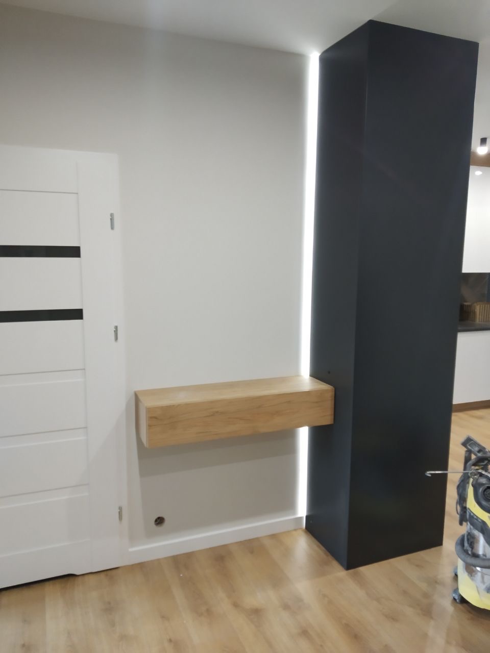 Nowoczesne wnętrze: biała ściana z drewnianą półką i białą szafą wnękową obok czarnej kolumny z oświetleniem LED, parkiet na podłodze.