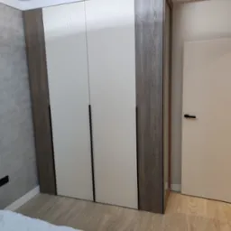 Nowoczesna szafa wnękowa z białymi, ryflowanymi frontami i obudową w kolorze ciemnego drewna, widoczna z perspektywy ł&oacute;żka w sypialni.