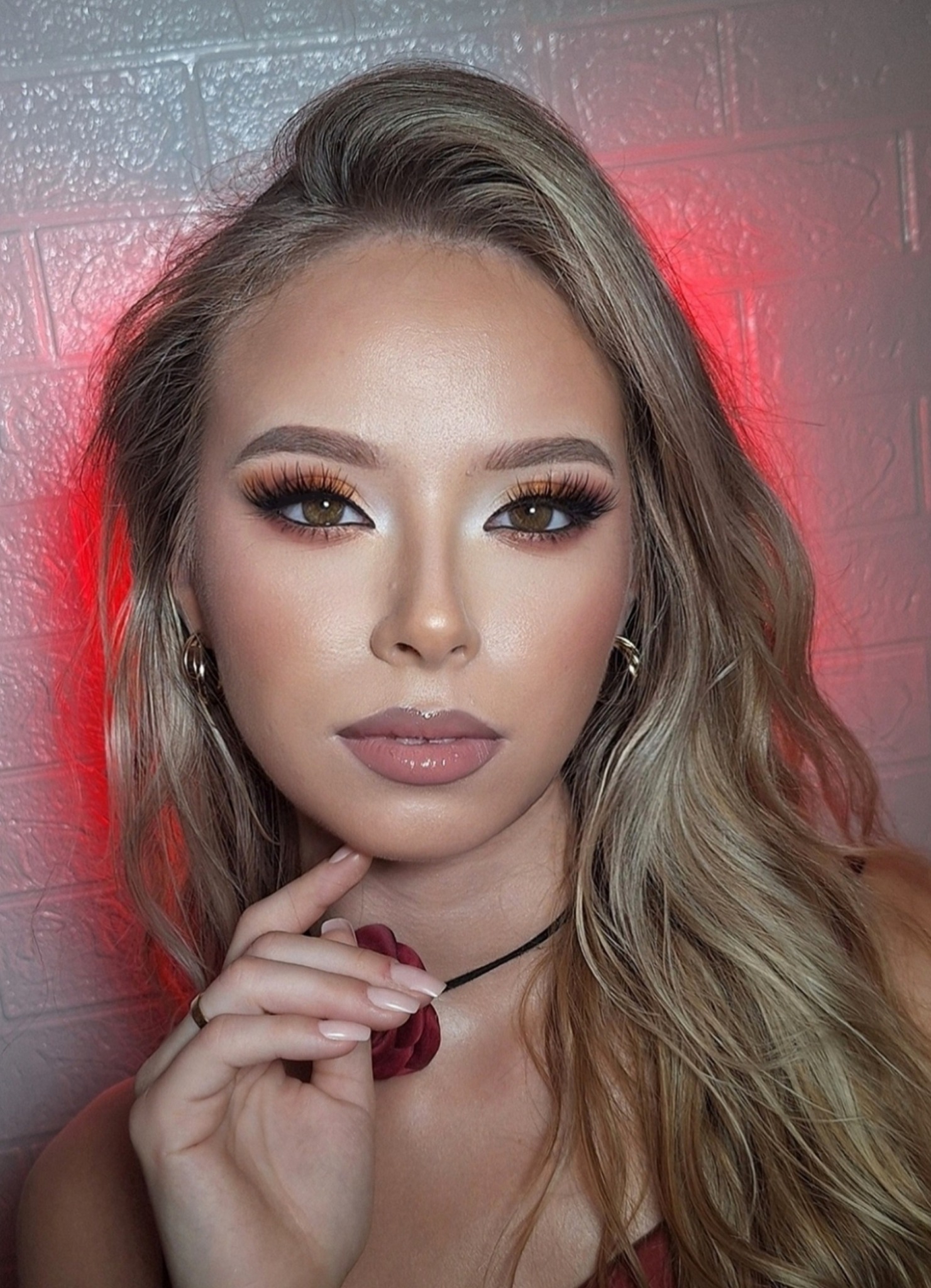 Portret kobiety z profesjonalnym makijażem oczu w odcieniach brązu i beżu, z długimi rzęsami i błyszczącymi ustami. Tło to ceglana ściana z czerwonym oświetleniem.