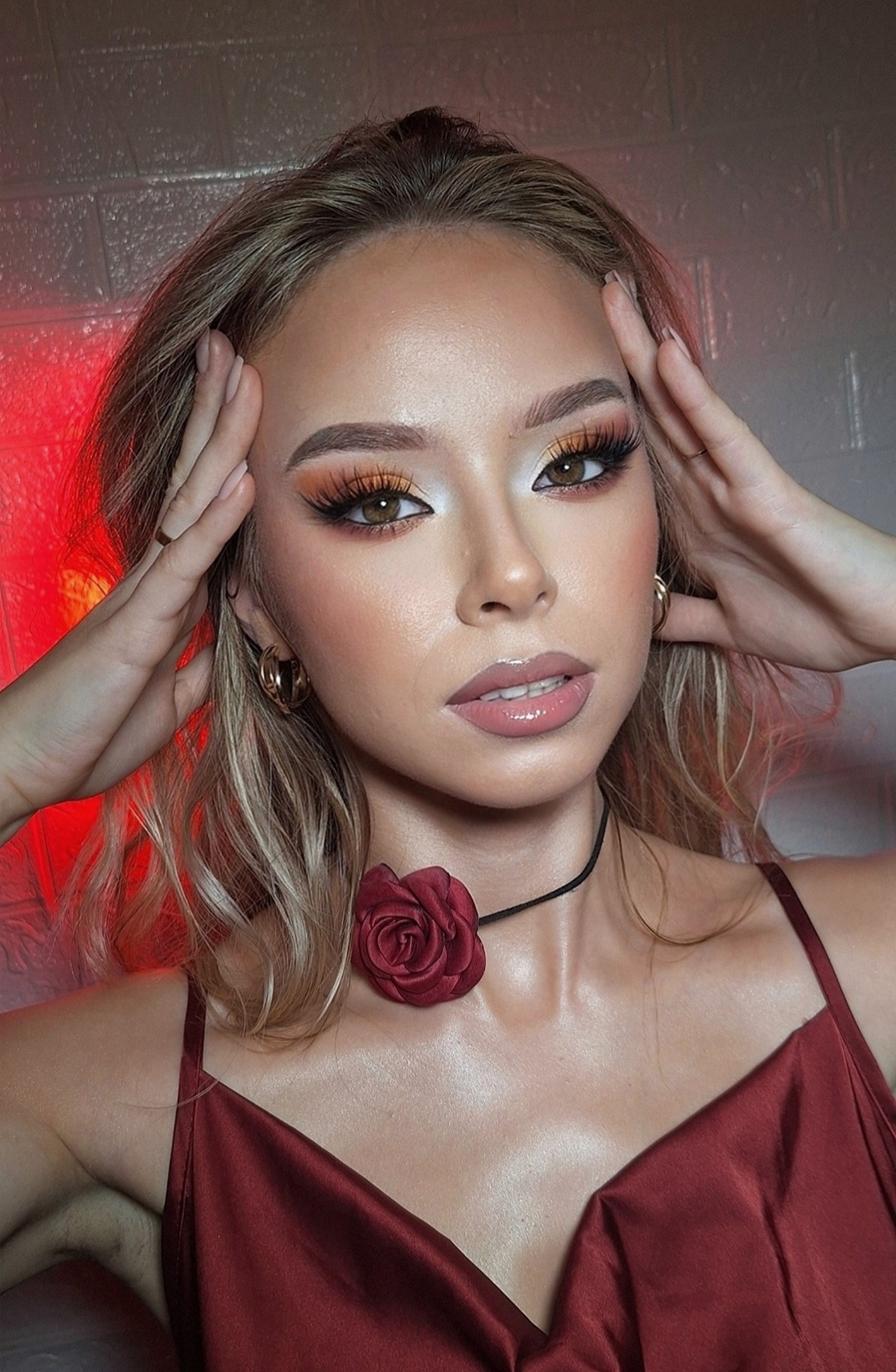 Portret kobiety z wyrazistym makijażem oczu w odcieniach brązu i złota, choker z różą i satynowa sukienka na tle ceglanej ściany z czerwonym oświetleniem.