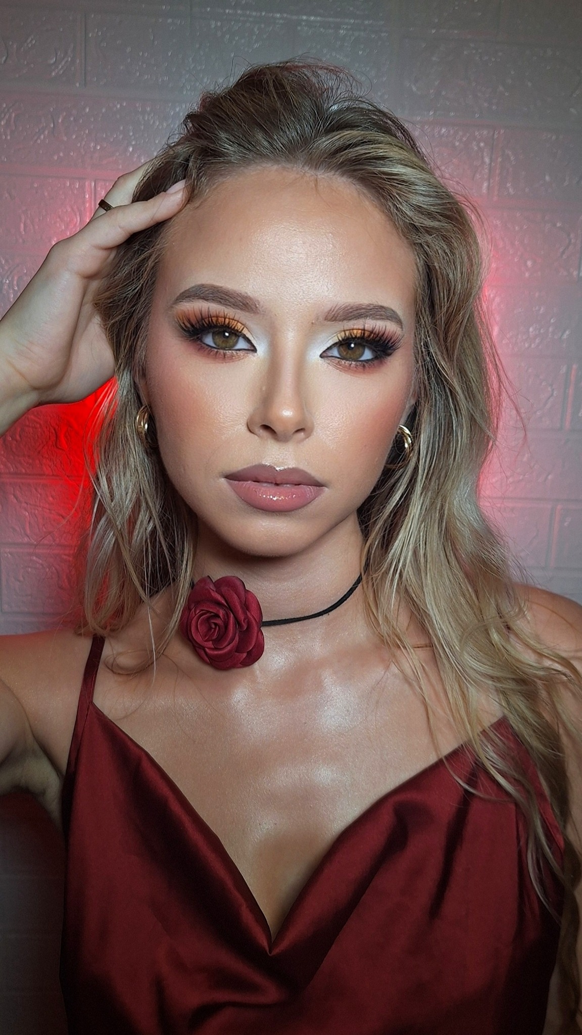 Portret kobiety z profesjonalnym makijażem oczu w odcieniach brązu i złota, chokerem z czerwoną różą i satynową sukienką na tle ceglanej ściany.