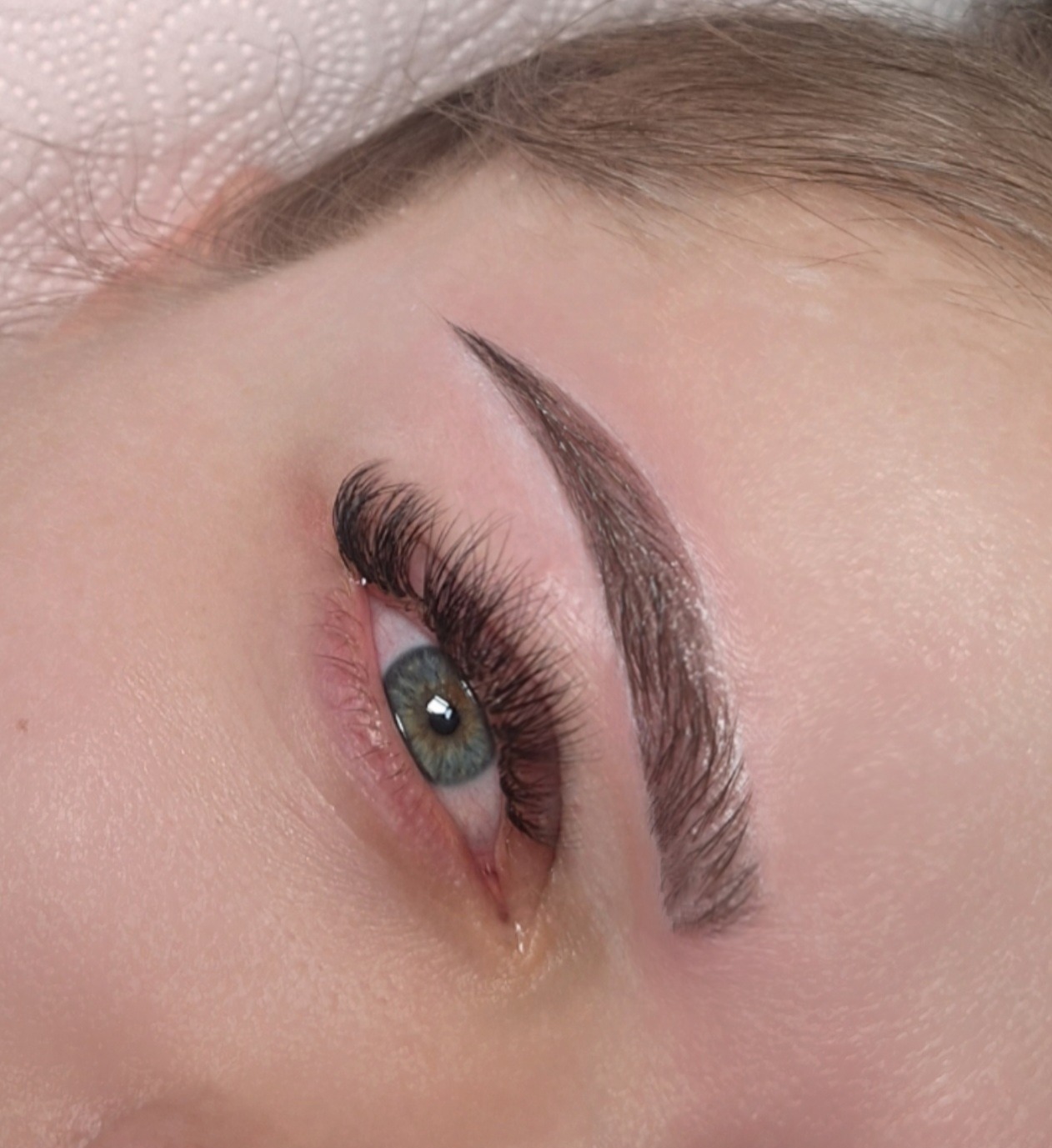 Zbliżenie na oko z przedłużonymi rzęsami i wyregulowanymi brwiami. Widoczna tęczówka oka w odcieniach zieleni i brązu. Delikatny makijaż podkreśla naturalne piękno.