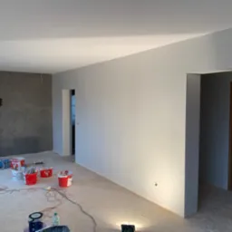 Wnętrze pokoju w trakcie prac: pomalowane na szaro ściany, widoczne wiaderka z farbą, pędzle, kabel, oraz surowa podłoga przed położeniem paneli.