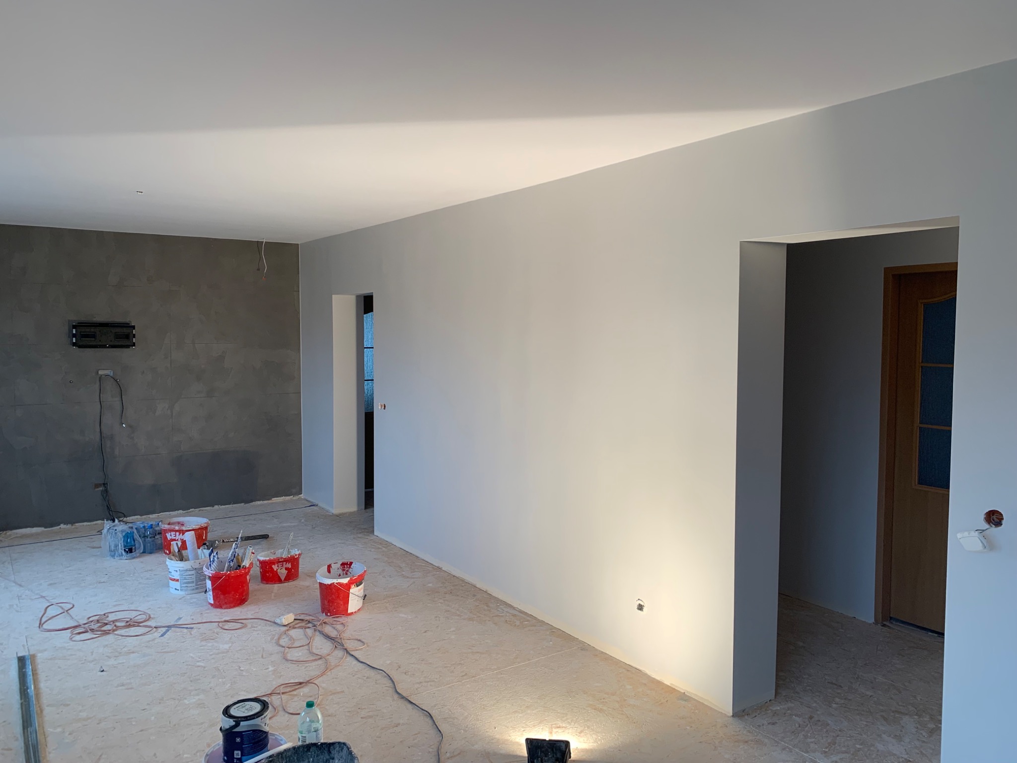 Wnętrze pokoju w trakcie prac: pomalowane na szaro ściany, widoczne wiaderka z farbą, pędzle, kabel, oraz surowa podłoga przed położeniem paneli.