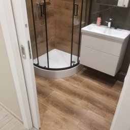 Jacek Wróbel - Nowoczesna łazienka z kabiną prysznicową narożną z czarnymi profilami, białą umywalką z szafką podwieszaną i płytkami imitującymi drewno na podłodze i ścianach.