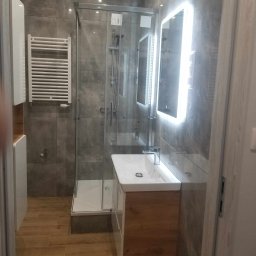 Jacek Wróbel - Nowoczesna łazienka z kabiną prysznicową, grzejnikiem drabinkowym i podświetlanym lustrem, wykończona szarymi płytkami i drewnopodobną podłogą.