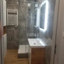 Nowoczesna łazienka z kabiną prysznicową, grzejnikiem drabinkowym i podświetlanym lustrem, wykończona szarymi płytkami i drewnopodobną podłogą.