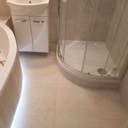 Jacek Wróbel - Jasna łazienka po remoncie z kabiną prysznicową, umywalką z szafką i fragmentem wanny, wykończona beżowymi płytkami.