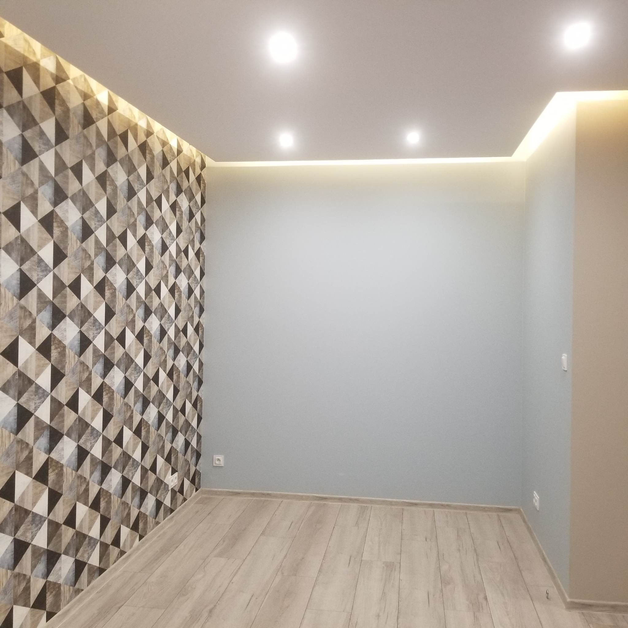 Pokój po remoncie z geometryczną tapetą na jednej ścianie, szarą farbą na pozostałych, drewnianą podłogą i oświetleniem LED pod sufitem.