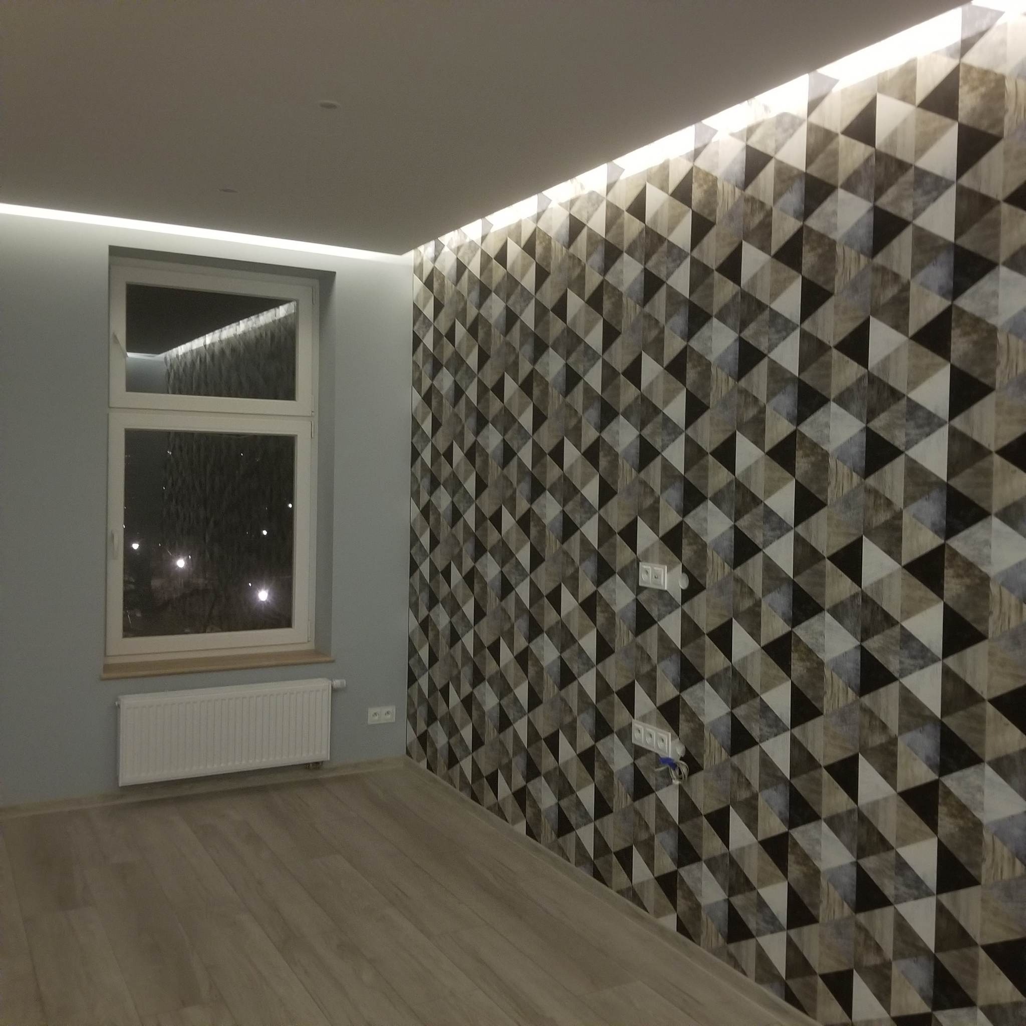 Pokój po remoncie z tapetą w trójkąty, podświetlonym sufitem i drewnianą podłogą; widoczne gniazdka elektryczne i grzejnik.