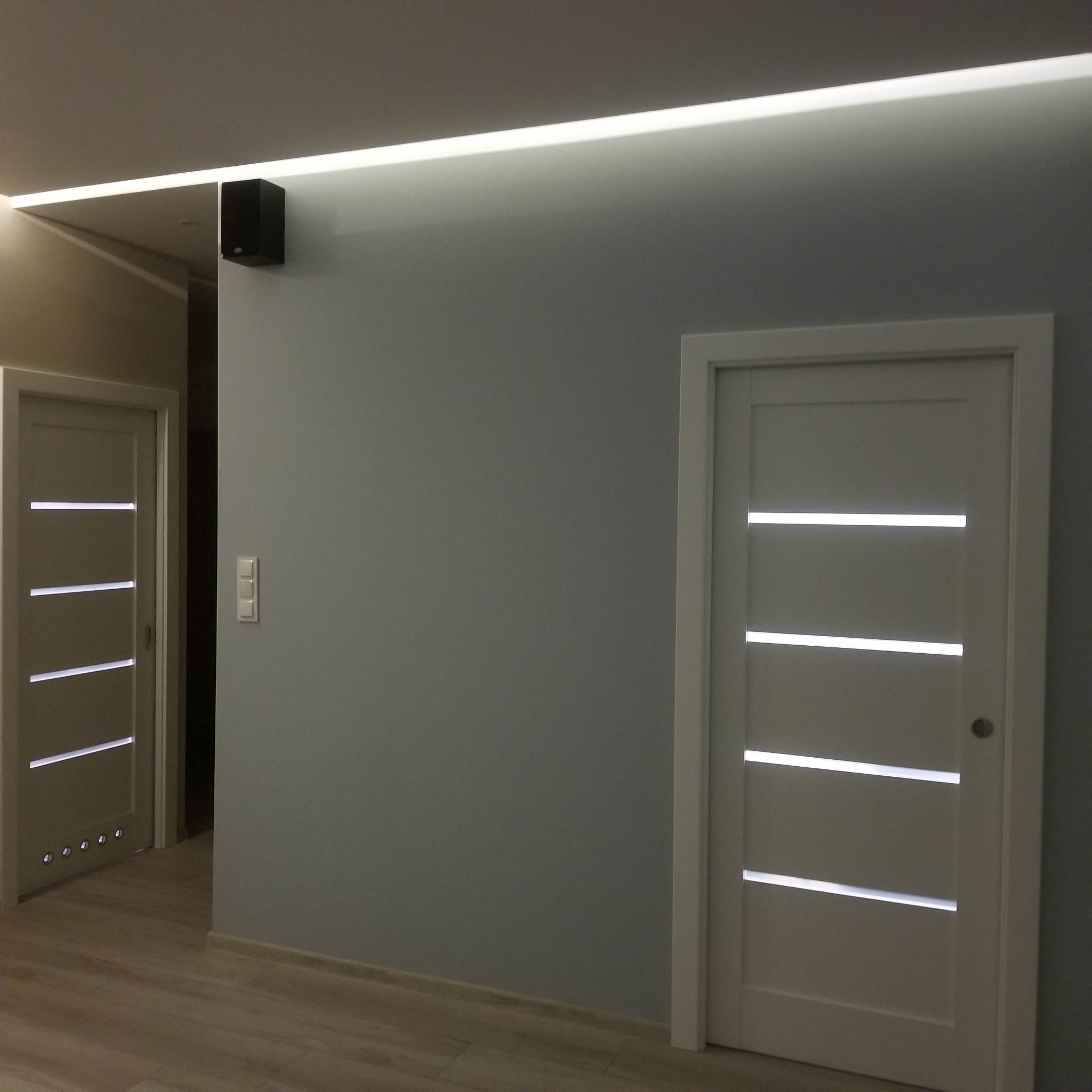 Szare wnętrze z dwiema białymi, nowoczesnymi drzwiami z poziomymi przeszkleniami, podświetlony sufit LED, gładkie ściany i jasna podłoga.