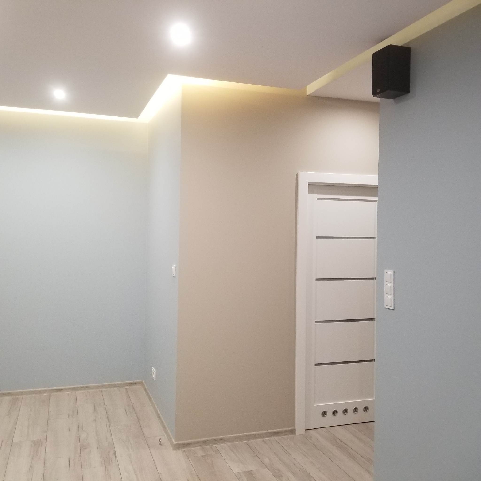 Nowoczesne wnętrze po remoncie: jasne ściany w odcieniach szarości i beżu, podświetlany sufit podwieszany z oświetleniem LED, białe drzwi z poziomymi frezami i drewniana podłoga.