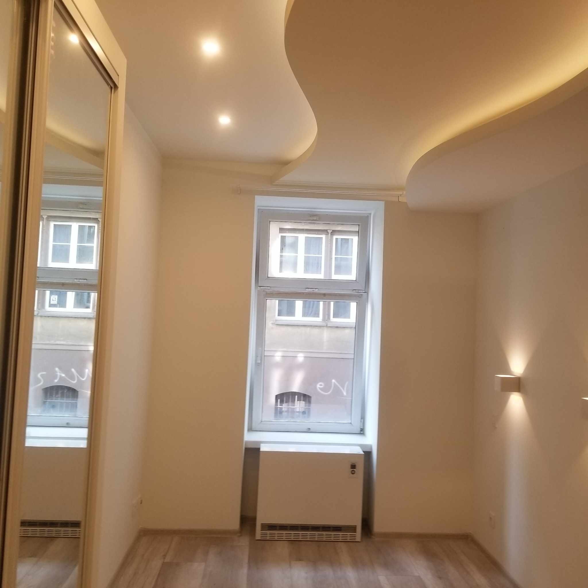 Jasne wnętrze po remoncie z falistym sufitem podwieszanym z oświetleniem LED, parkietem, grzejnikiem pod oknem i lustrem w szafie wnękowej.