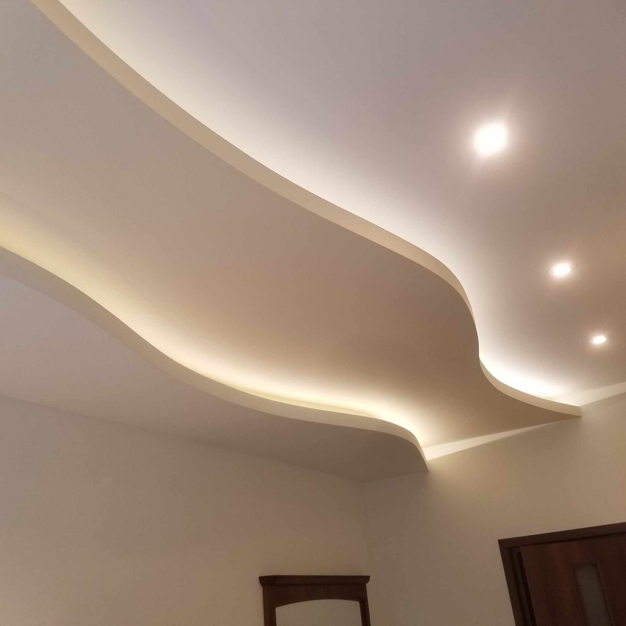 Wielopoziomowy, podwieszany sufit w kształcie fali, oświetlony taśmami LED, z trzema okrągłymi lampami punktowymi i widocznym fragmentem ściany z drzwiami.