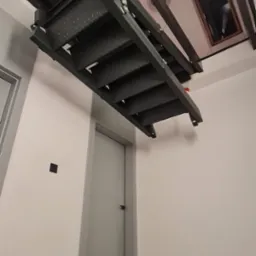 Zamknięte, składane schody strychowe z metalową konstrukcją i perforowanymi stopniami, widoczne od dołu, zamontowane w stropie pomieszczenia z drzwiami i oknem.