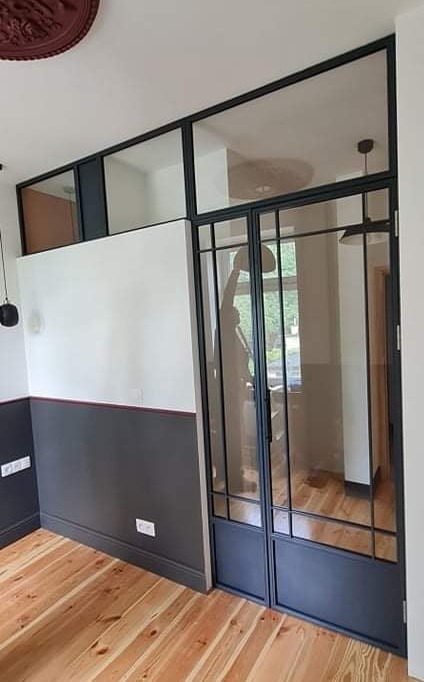 Wnętrze z drewnianą podłogą i ścianami pomalowanymi na biało i szaro, oddzielone od sąsiedniego pomieszczenia szklaną ścianą z czarnymi metalowymi ramami w stylu industrialnym.