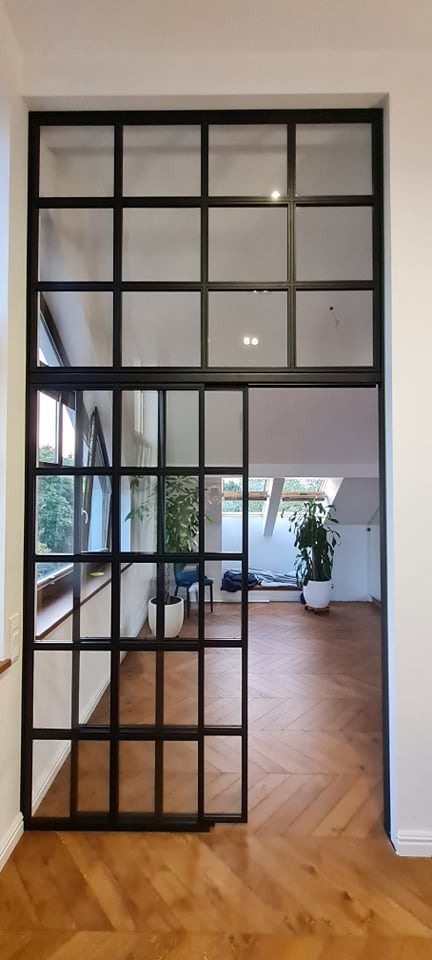 Przeszklona, przesuwna ściana działowa z czarnymi profilami, oddzielająca pokój na poddaszu z parkietem ułożonym w jodełkę.