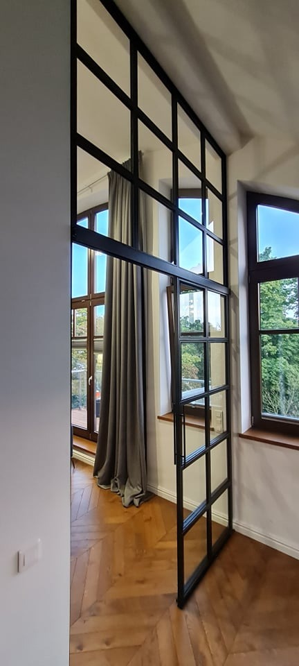 Loftowa ścianka działowa z czarnej stali i szkła, z przesuwnymi drzwiami, dzieląca przestrzeń w jasnym wnętrzu z parkietem w jodełkę i oknami z widokiem na zieleń.