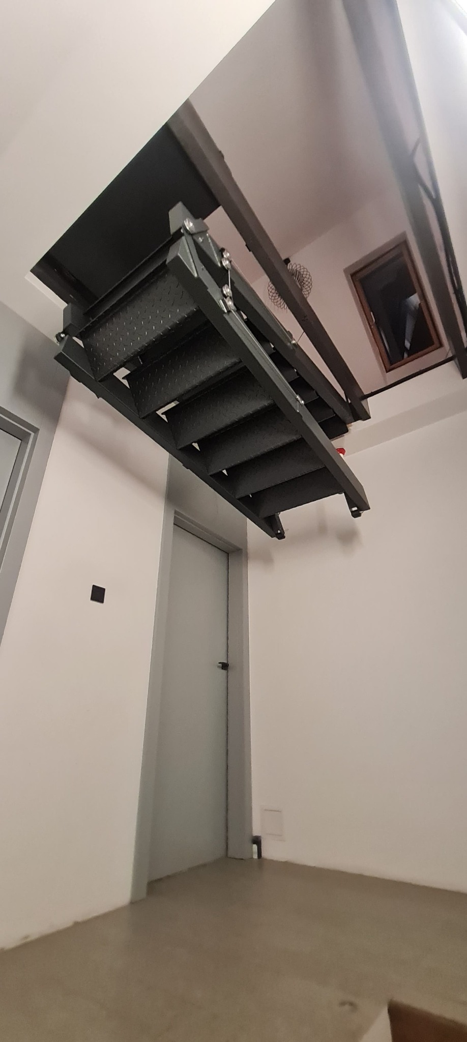 Zamknięte, składane schody strychowe z metalową konstrukcją i perforowanymi stopniami, widoczne od dołu, zamontowane w stropie pomieszczenia z drzwiami i oknem.