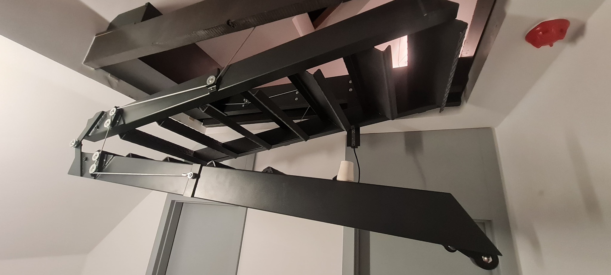 Składana konstrukcja stalowa podwieszona do sufitu, prawdopodobnie element hali lub magazynu, widoczne elementy mocujące i mechanizm składania.