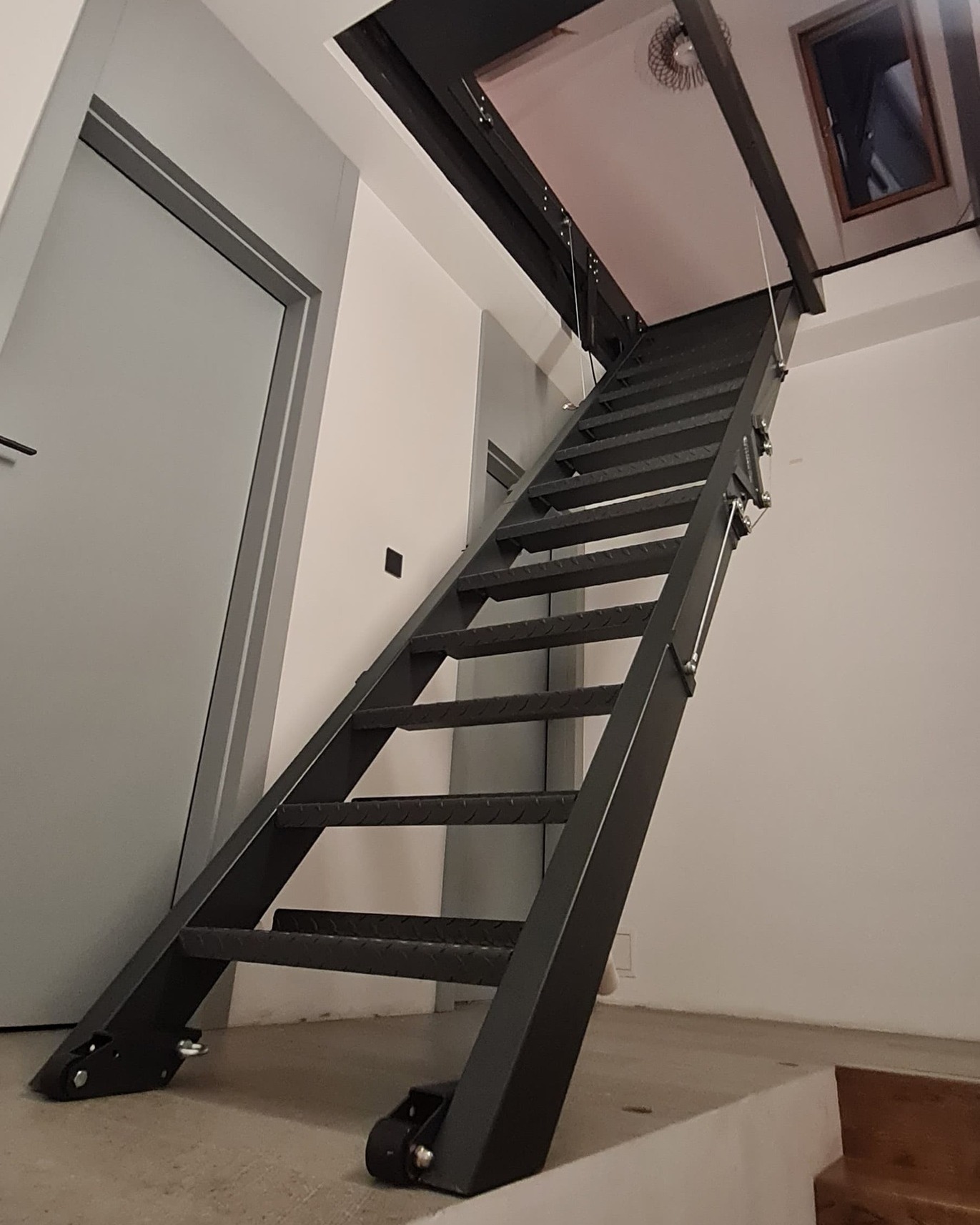 Metalowe schody strychowe składane, z widocznymi elementami mocującymi do sufitu i podłogi, w minimalistycznym wnętrzu z białą ścianą i szarą ościeżnicą drzwiową.