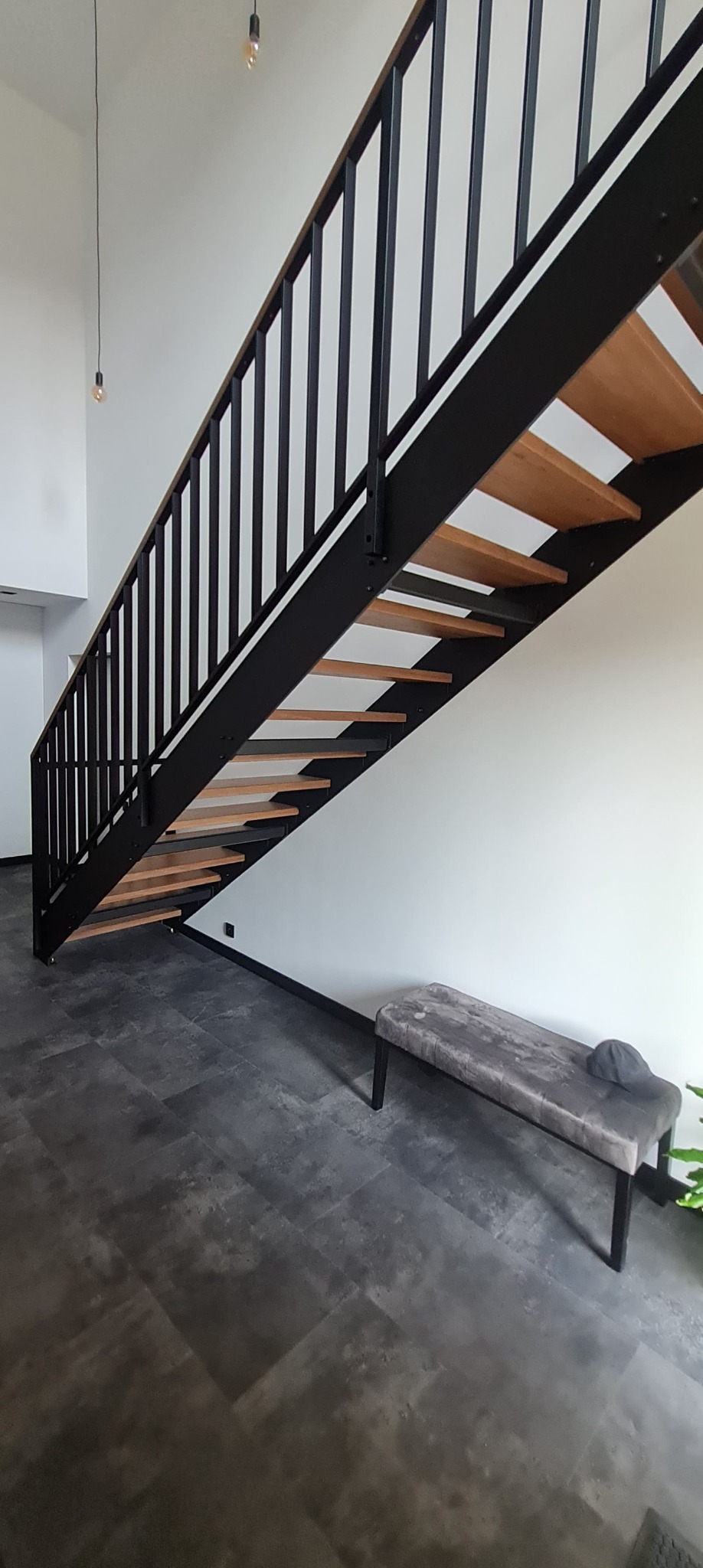 Nowoczesne schody z drewnianymi stopniami i czarną metalową konstrukcją w minimalistycznym wnętrzu z szarą podłogą i ławką.