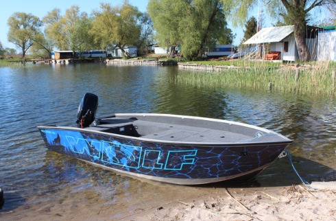 Aluminiowa łódź motorowa z czarnym silnikiem i niebieskim wzorem 'WOLF' zacumowana na piaszczystym brzegu jeziora, w tle widoczne domki letniskowe.