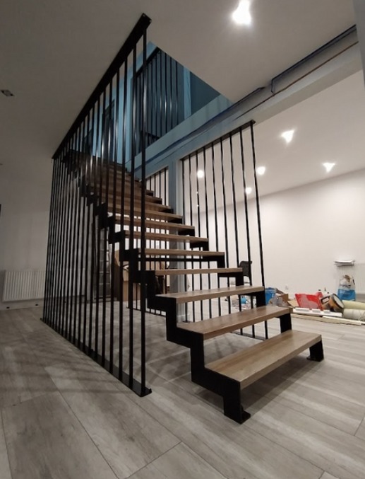 Nowoczesne schody wewnętrzne z drewnianymi stopniami i czarną, minimalistyczną balustradą z pionowych prętów, w jasnym wnętrzu z panelami podłogowymi imitującymi drewno.