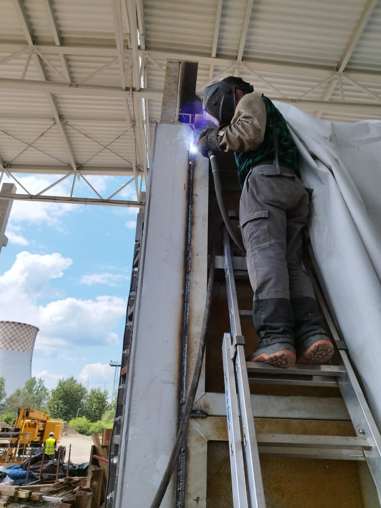 Spawacz w masce na drabinie spawa pionowy element konstrukcyjny w hali, widoczne iskry i ślady spawania, w tle komin chłodni i podnośnik koszowy.