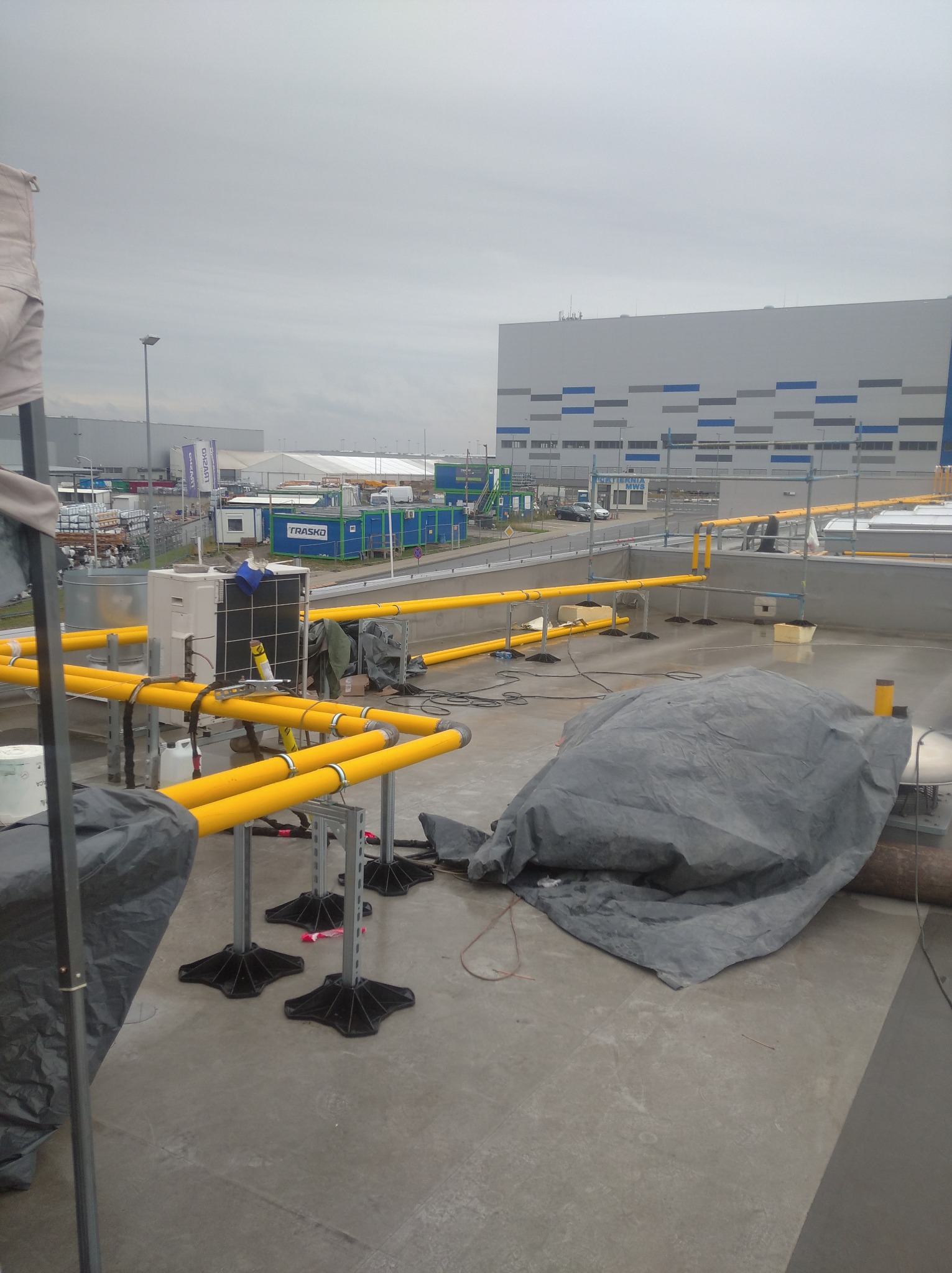 Instalacja żółtych rur na dachu przemysłowego budynku, widoczne podesty, klimatyzator i elementy pod plandeką.
