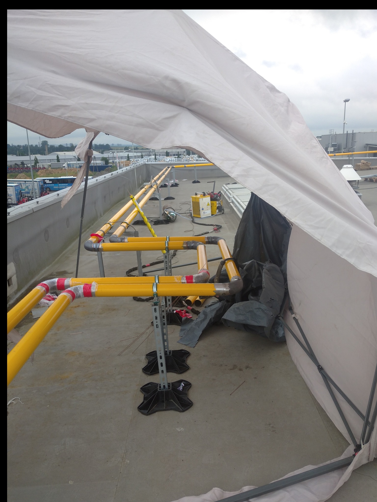 Żółte rury połączone kolankami z czerwoną taśmą, ustawione na metalowych wspornikach, z osłoną z białego materiału i widocznym sprzętem spawalniczym w tle, na betonowym podłożu.