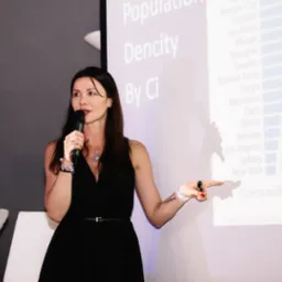 Kobieta w czarnej sukience prezentuje dane o gęstości zaludnienia miast na ekranie projektora, trzymając mikrofon w lewej ręce i wskazując na ekran prawą ręką z czerwonym lakierem na paznokciach.