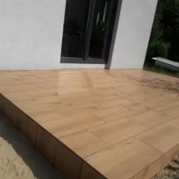 Wykończony taras z płytek imitujących drewno, przylegający do białego budynku z czarnym oknem, obok piasek i wanna.