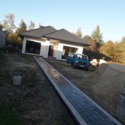 Łukasza Wojtak - Nowo ułożony, prosty chodnik z ciemnej kostki brukowej z jasnoszarymi obrzeżami prowadzi do nowoczesnego, parterowego domu z ciemnym dachem; w tle zaparkowany turkusowy bus i usypana pryzma piasku.