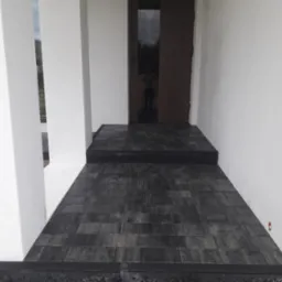 Ciemnoszara kostka brukowa prowadząca do wejścia domu z białymi filarami i drewnianymi drzwiami z odblaskiem sylwetki.