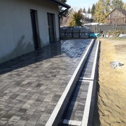 Łukasza Wojtak - Ułożona, grafitowa kostka brukowa z białymi obrzeżami na tarasie przylegającym do budynku, z widoczną stertą nieużytych kostek w tle.