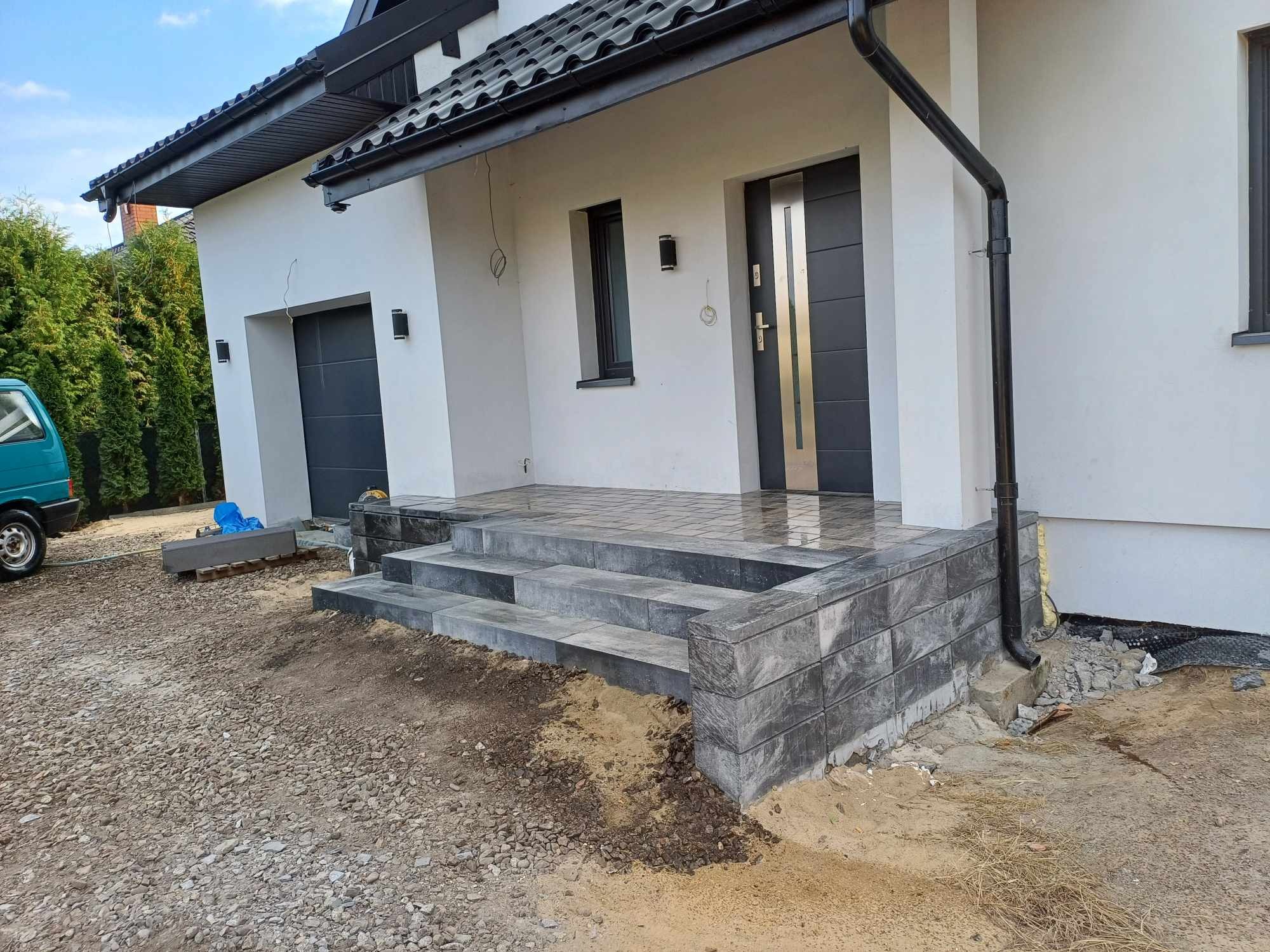 Nowe, szare schody wejściowe z kostki brukowej prowadzące do domu z białą elewacją i czarnymi detalami, widoczne pozostałości piasku po pracach montażowych.
