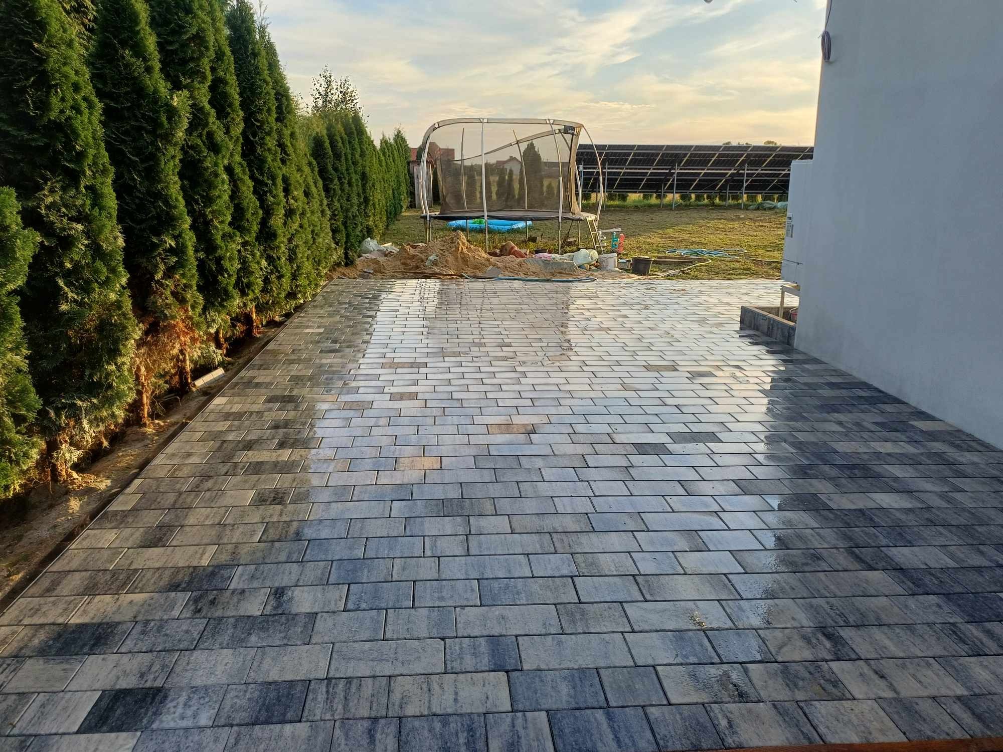 Świeżo ułożona, mokra kostka brukowa w odcieniach szarości, przylegająca do ściany budynku i rzędu wysokich tuj, z trampoliną i panelami słonecznymi w tle.