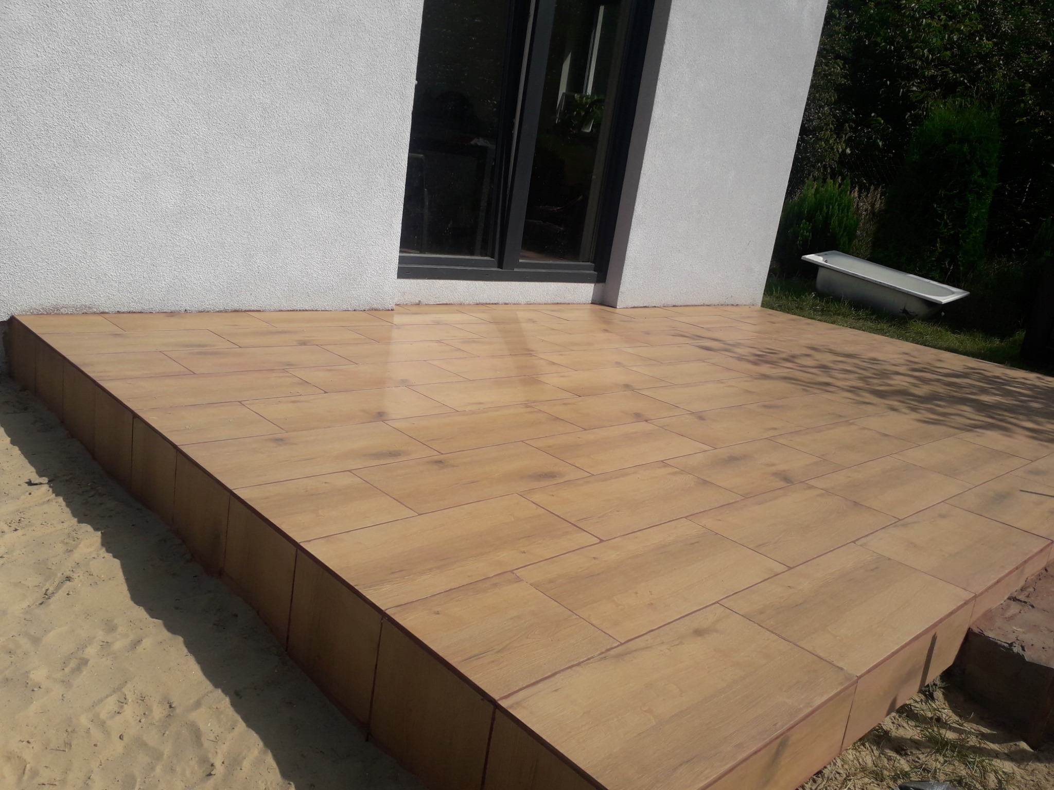 Wykończony taras z płytek imitujących drewno, przylegający do białego budynku z czarnym oknem, obok piasek i wanna.
