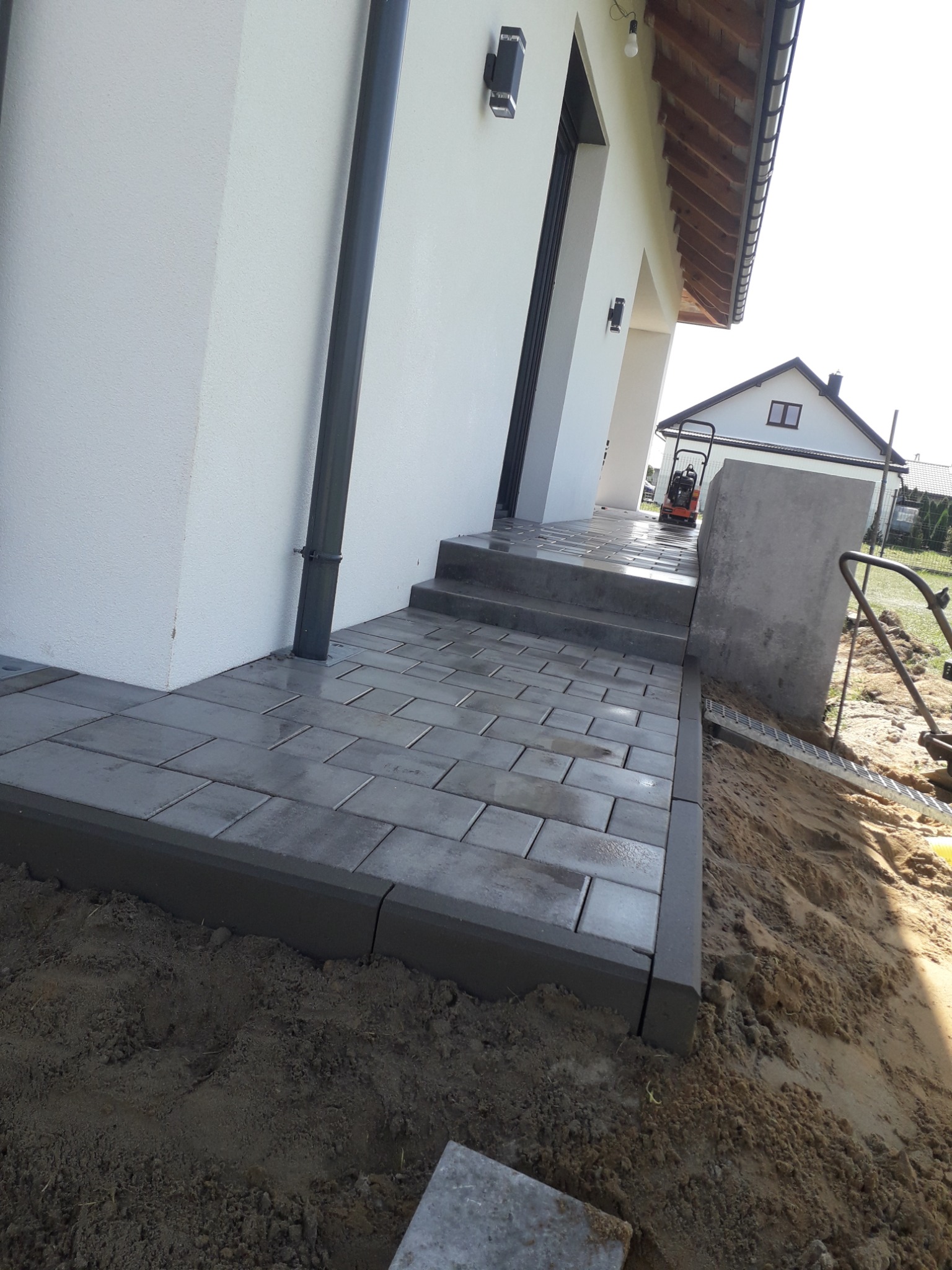 Ułożona szara kostka brukowa prowadząca do wejścia domu z widoczną ubijarką w tle, obrzeża kostki z ciemnoszarego betonu.