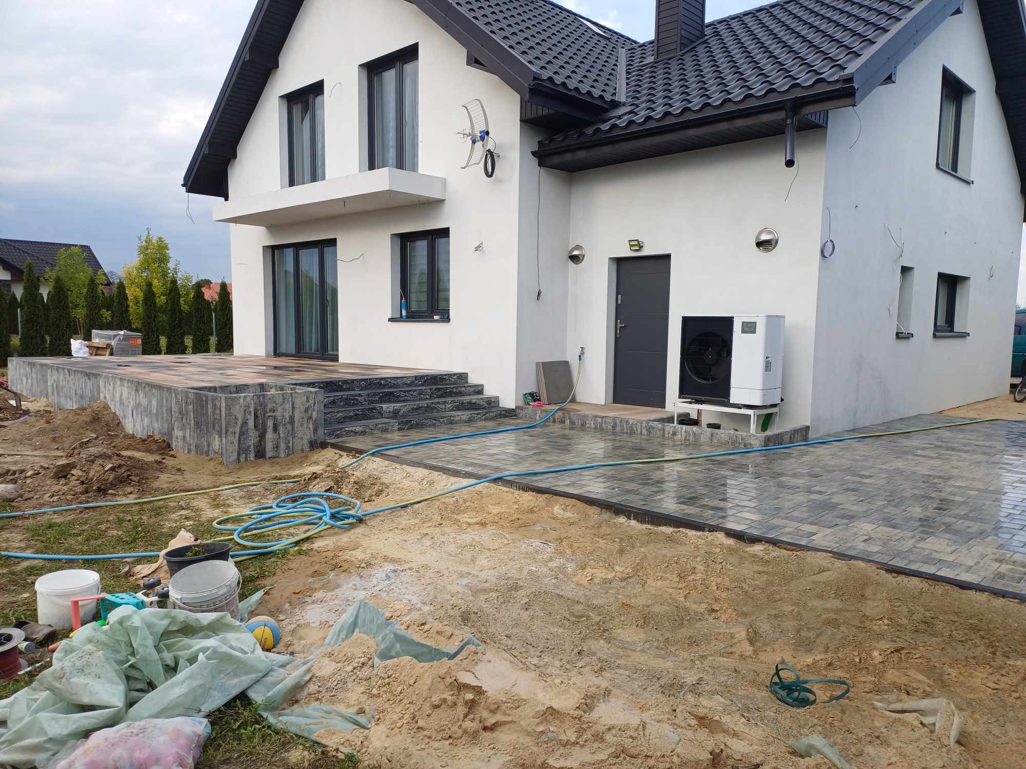Nowoczesny dom z szarą kostką brukową w trakcie układania, widoczne narzędzia i piasek, elewacja biała z czarnymi ramami okiennymi, pompa ciepła na zewnątrz.