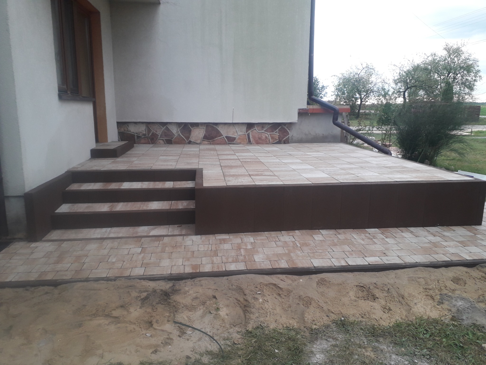 Ułożony taras z jasnej kostki brukowej z brązowymi obrzeżami i schodami przy ścianie budynku z kamienną podmurówką, widok z poziomu gruntu.